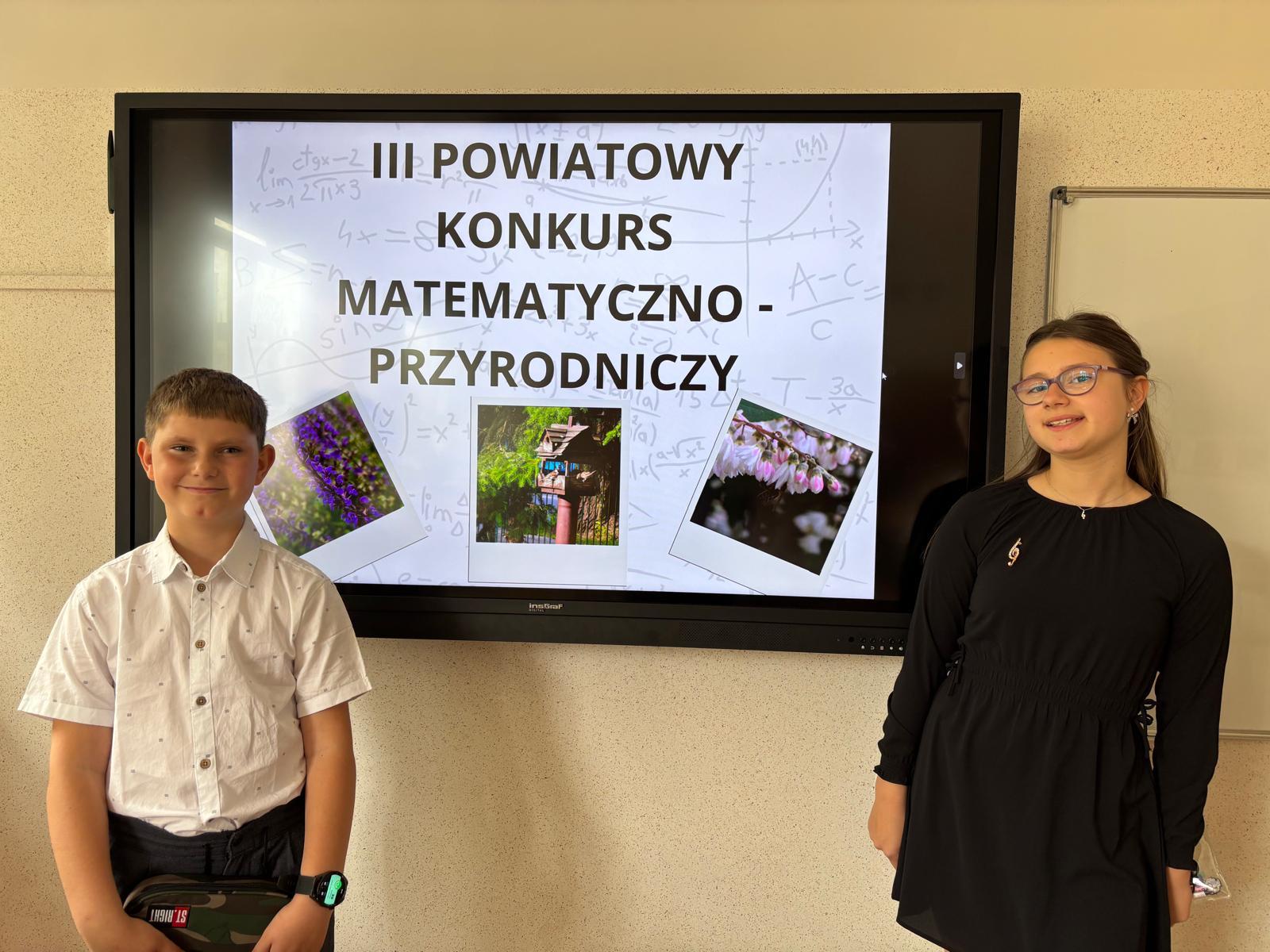 Chlopiec i dziewczynka na tle nazwy "III Powiatowy Konkurs matematyczno-przyrodniczy" wyświetlanej na ekranie